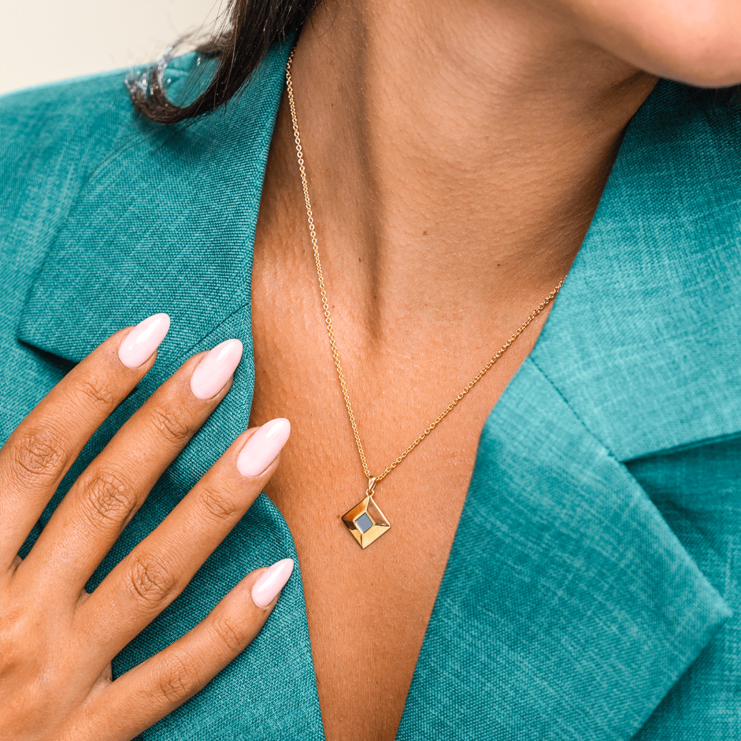 A person wearing a gold necklace with a geometric pendant and a teal blazer.